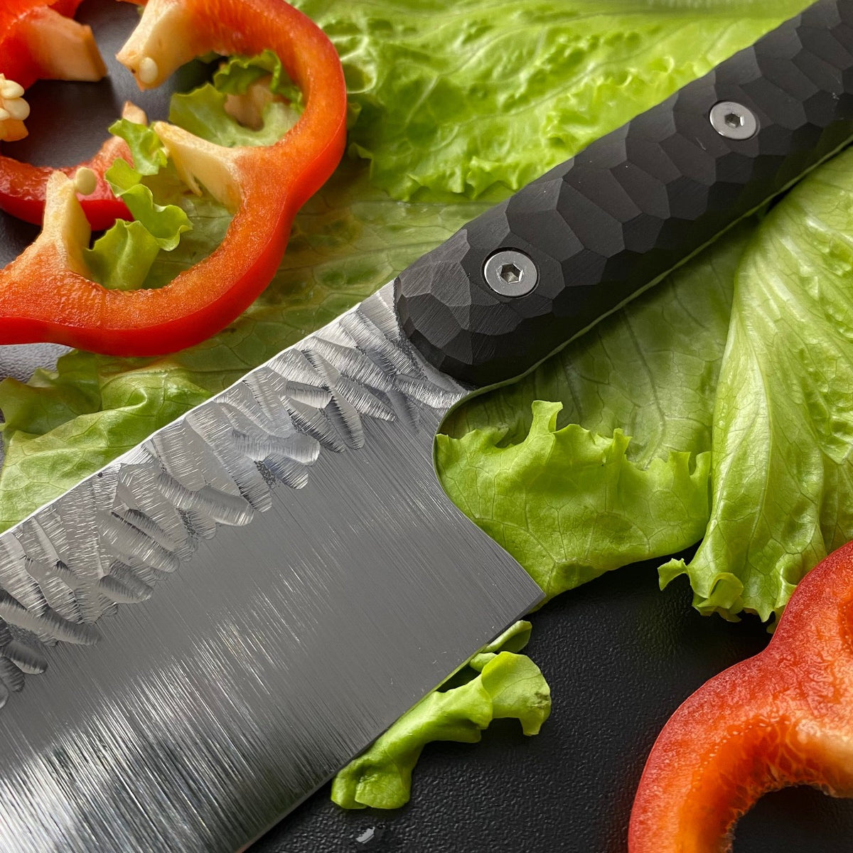 Wide stainless steel santoku knife on dark board with sliced red bell pepper and green lettuce