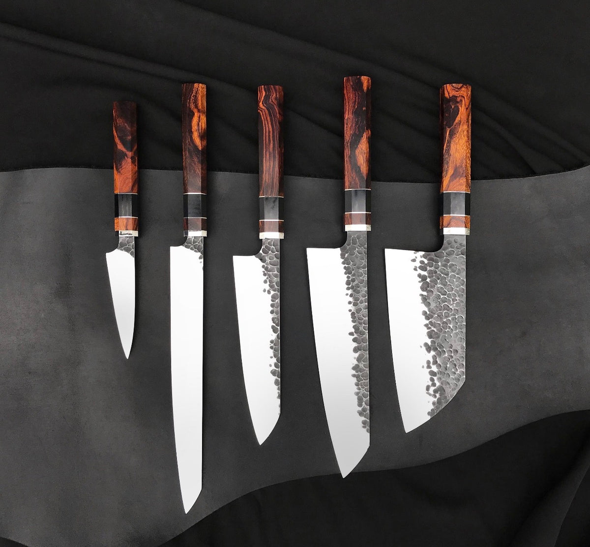 Chef kitchen knife set of five handmade knives with hammered blades and wood handles on black fabric
