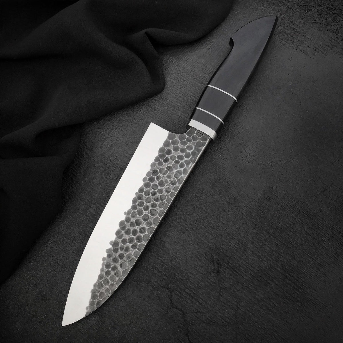 Hammered kitchen chef knife with polished edge and black Micarta handle on dark surface

