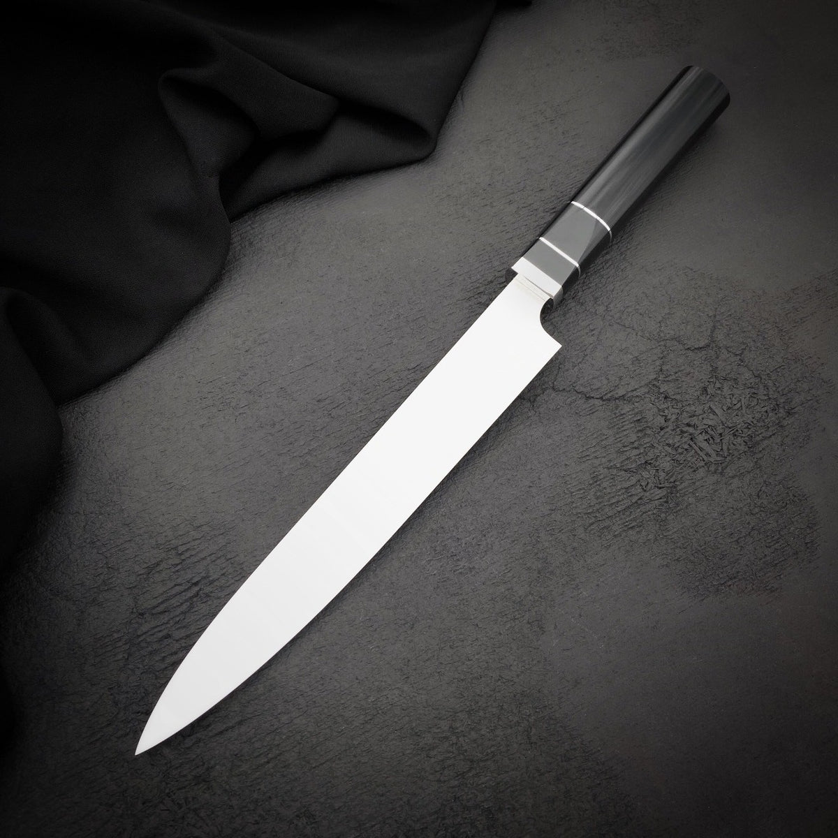 Handmade 9 inch sushi slicer with straight spine and pointed tip beside black fabric
