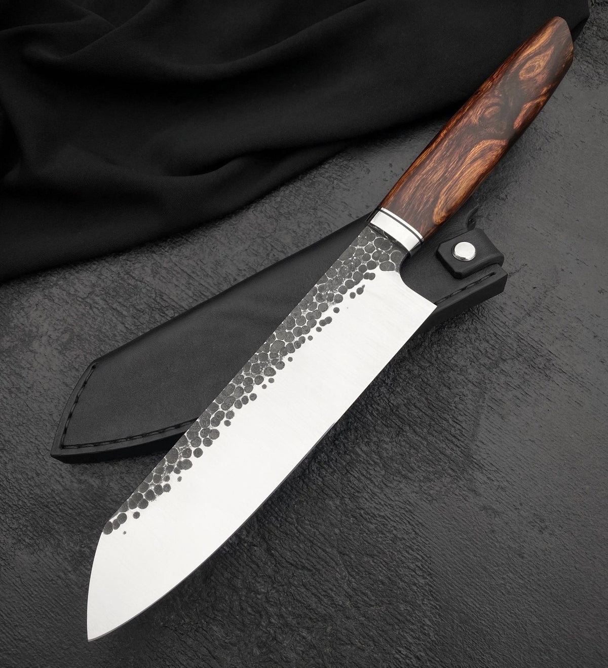 Chef knife with sheath showing hammered santoku blade and swirled brown wood handle on dark surface
