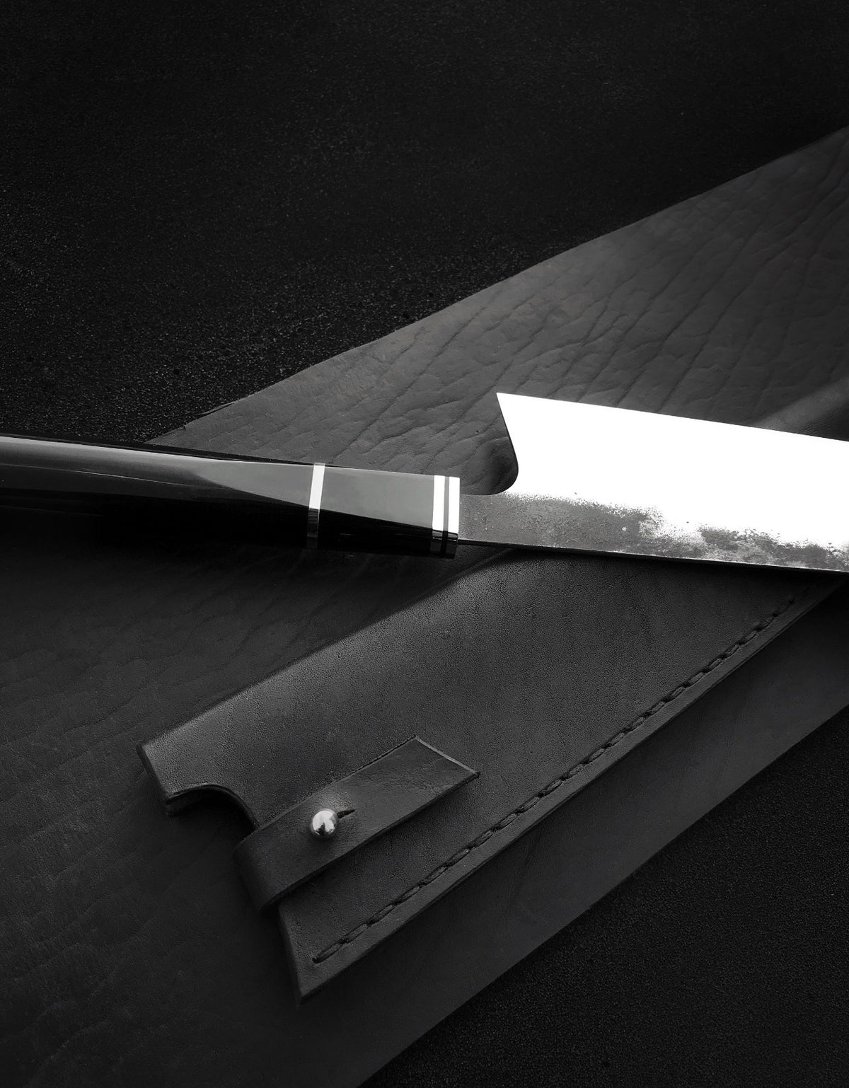 Handmade chef knife with angled kiritsuke tip lying next to stitched black leather sheath
