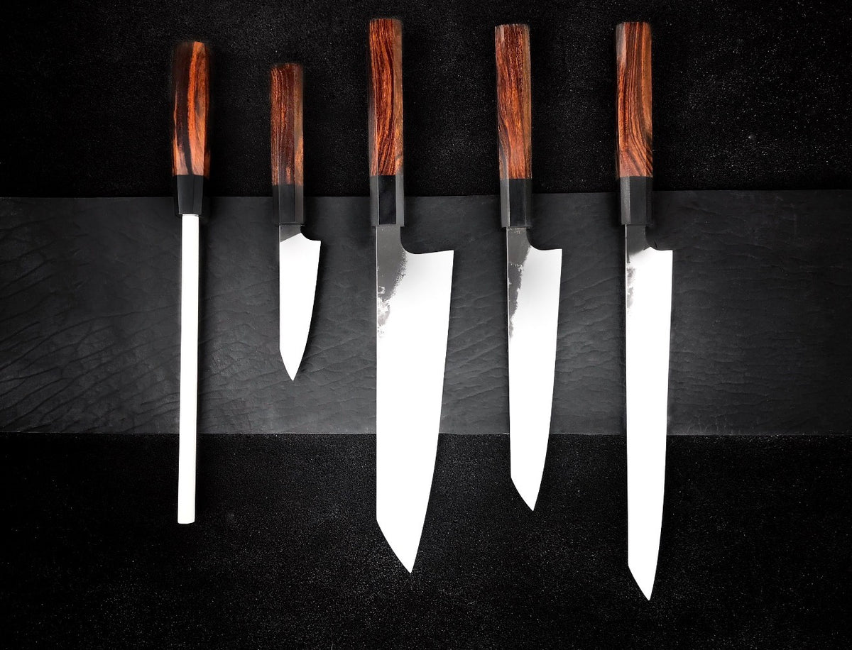 Wood handle knife set with four kitchen knives and a honing rod laid out in a row on black
