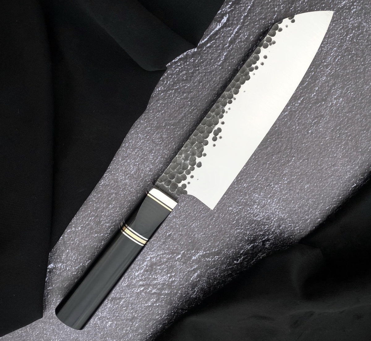 Santoku blade kitchen cooking knife with hammered 440C blade and black Micarta handle on gray textured surface
