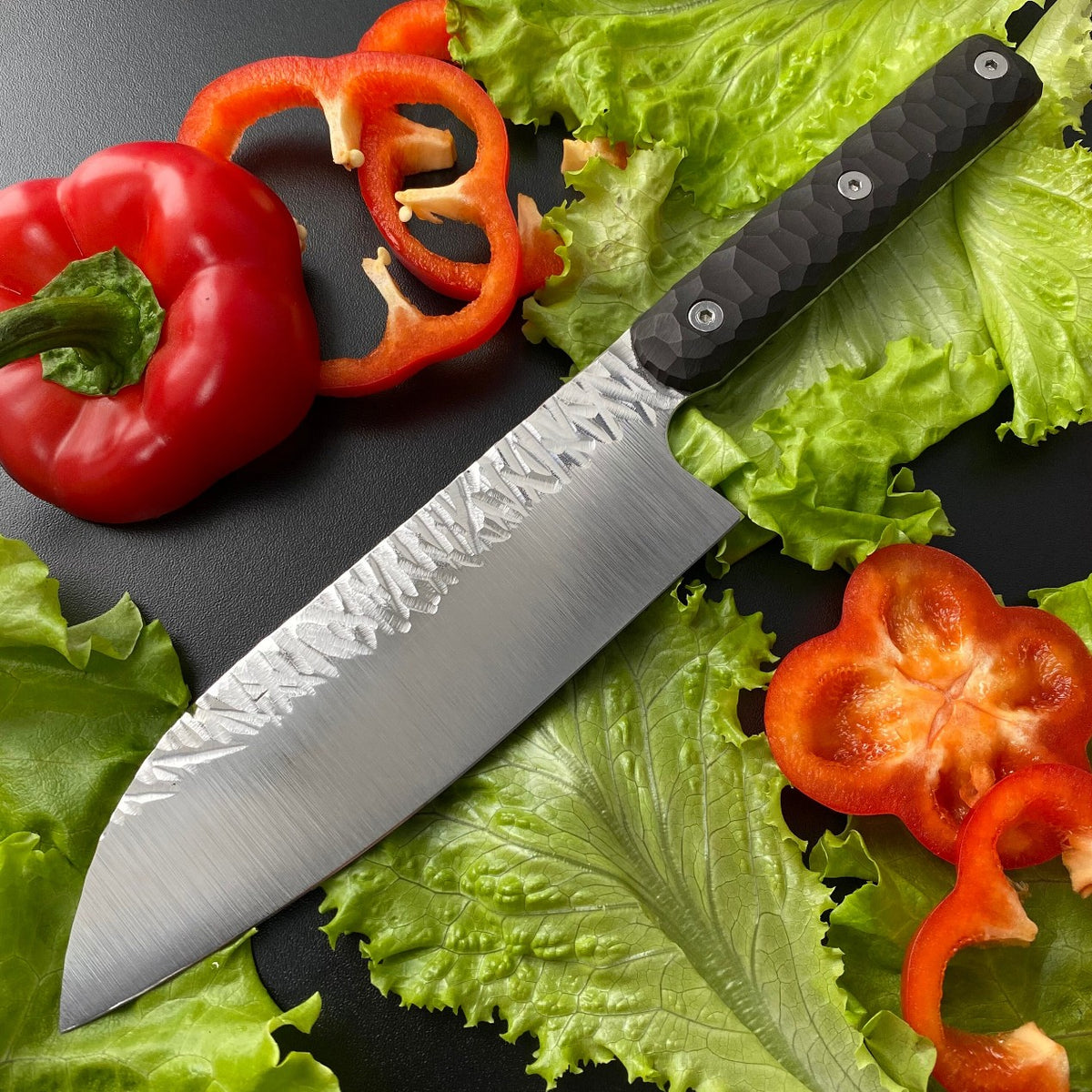 Santoku chef knife with hammered 440C blade and black textured G10 handle among red peppers and lettuce
